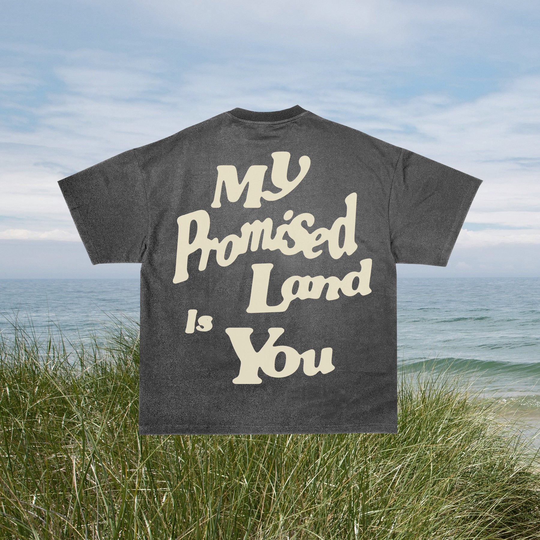 A gray t-shirt with the text 'MY PROMISED LAND IS YOU' printed on the front, displayed on a hanger against a beach backdrop.