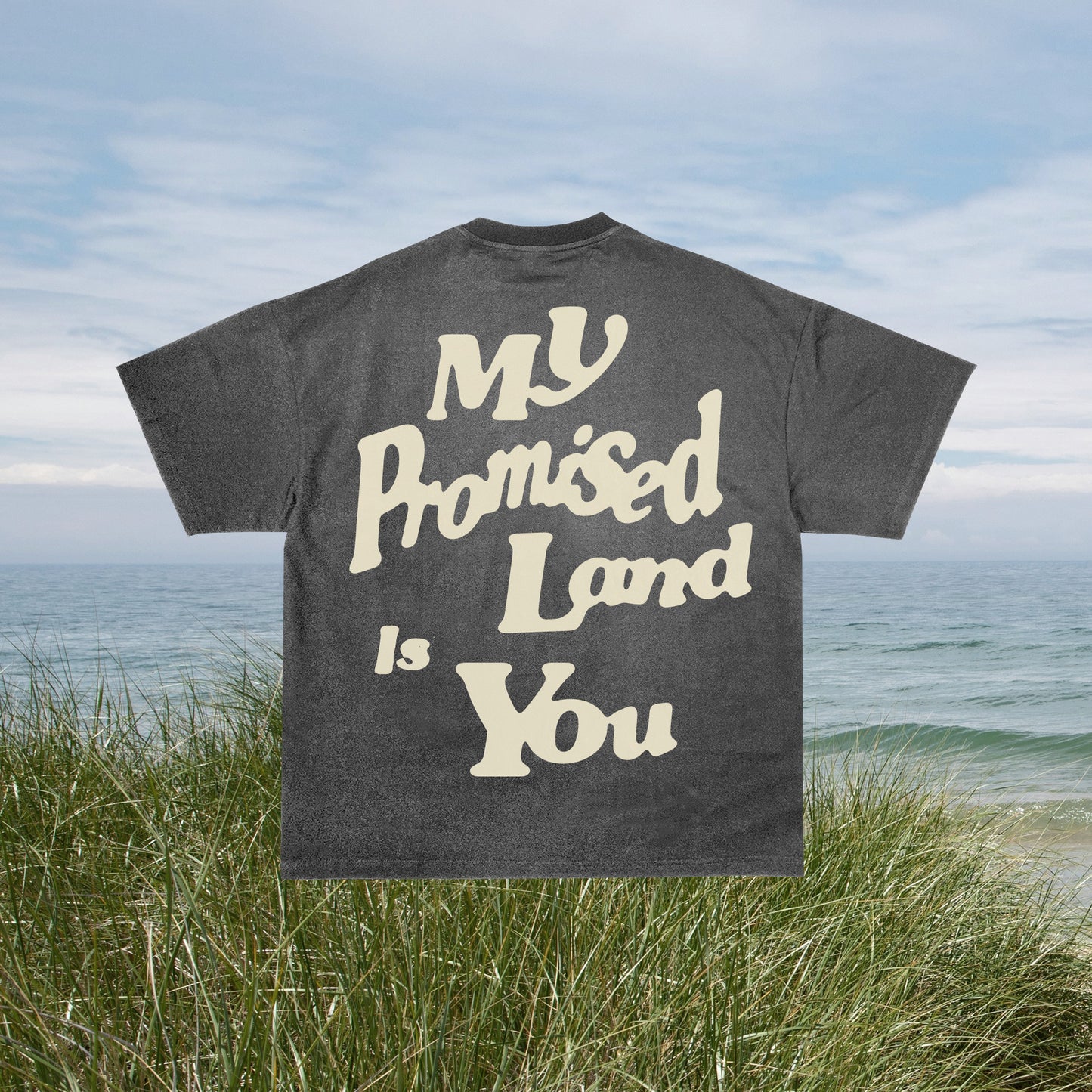 A gray t-shirt with the text 'MY PROMISED LAND IS YOU' printed on the front, displayed on a hanger against a beach backdrop.